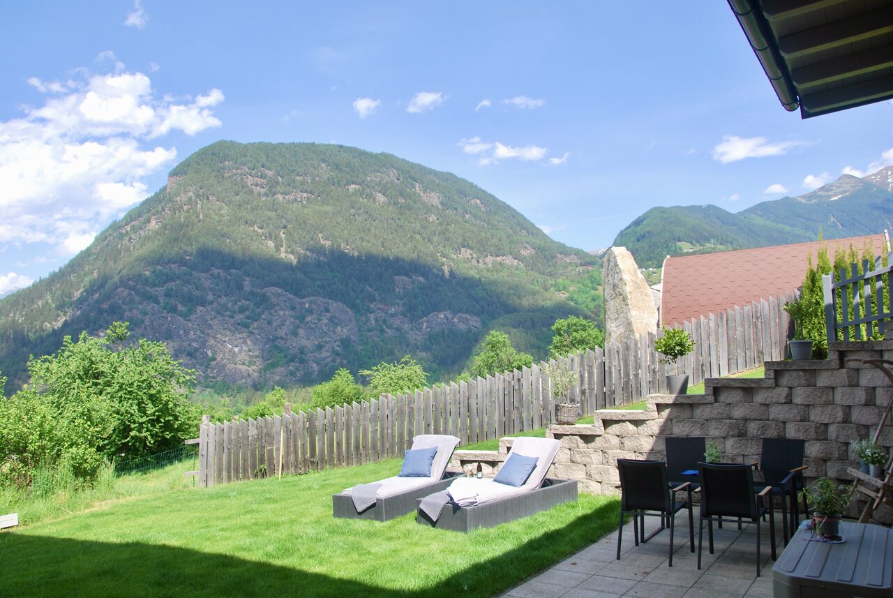 Blick aus dem Garten der Ferienwohnung auf den bewaldeten Berghang gegenüber, davor Liegestühle auf dem Rasen und eine Terrasse mit Tisch und Stühlen.